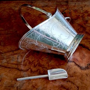 May include: A silver-toned metal ice bucket featuring an arched handle and intricate etched patterns on the exterior surface. The bucket is accompanied by a matching small metal scoop with a flat handle and a rounded, shovel-like head.