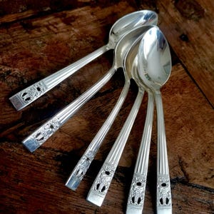 May include: Five silver-toned metal spoons arranged in a fan pattern. Each spoon features a polished oval bowl and a handle decorated with vertical fluted lines and a rectangular floral motif at the base of the stem near the end of the handle.