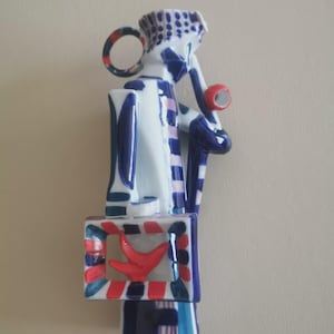 Puede incluir: Una figura de cerámica que representa a un personaje estilizado con un patrón geométrico en azul, blanco y rojo. El personaje sostiene un objeto circular rojo y un marco rectangular que contiene una forma de pájaro roja, con un detalle a cuadros.