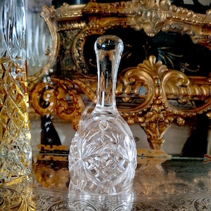 Puede incluir: Una campana de cristal tallado transparente con un patrón de diamantes, sobre una bandeja plateada. La campana tiene un mango largo y delgado y una parte superior redondeada. Un segundo objeto de vidrio es visible a la izquierda. El fondo presenta detalles dorados ornamentados.