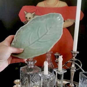 Peut inclure: Un plat en céramique en forme de feuille vert pâle est tenu devant une peinture et divers objets décoratifs. Le plat a une surface texturée et un motif de veine central. Autres articles : carafes en verre, bougeoirs et un plateau argenté.