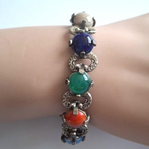 May include: A link bracelet featuring a series of round, smooth, multi-colored cabochon stones including beige, dark blue, green, and orange. The stones are set within ornate, textured silver-toned metal links that connect each circular element.