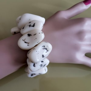 May include: A stretch bracelet composed of large, smooth, off-white pebble-shaped beads. Each bead features irregular black speckled markings across its surface. The beads are strung together in a continuous loop to form a chunky, rounded wrist accessory.