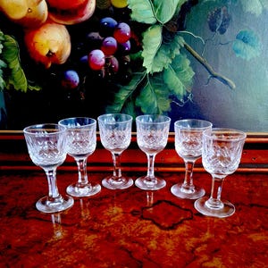 May include: Six clear crystal shot glasses with a diamond pattern. The glasses have a stemmed base and are arranged on a dark, glossy surface. A painting of fruit and leaves is in the background.