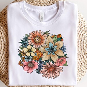 May include: White t-shirt featuring a colorful floral bouquet illustration. The bouquet includes various flowers in shades of orange, yellow, pink, and purple, with green leaves. The design is centered on the shirt, which is displayed folded on a woven surface.