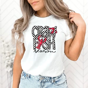 May include: White t-shirt with a black and white checkered design that says "Crawfish Season" with a red crawfish illustration.