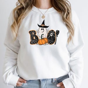 May include: White sweatshirt featuring a graphic of a ghost wearing a witch's hat and holding a pumpkin. The ghost is surrounded by a spiderweb design and the word "BOO" is written in black letters.