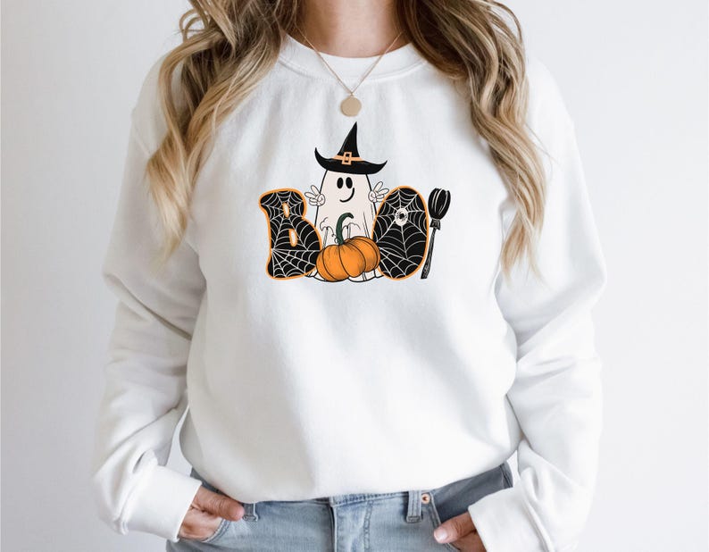 May include: White sweatshirt featuring a graphic of a ghost wearing a witch's hat and holding a pumpkin. The ghost is surrounded by a spiderweb design and the word "BOO" is written in black letters.