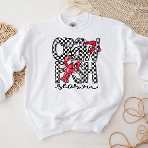 May include: White sweatshirt with a black and white checkered design that says "Crawfish Season" with a red crawfish illustration.