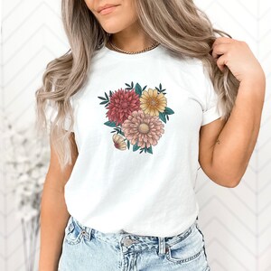 May include: White t-shirt with a floral print featuring pink, yellow, and red flowers.
