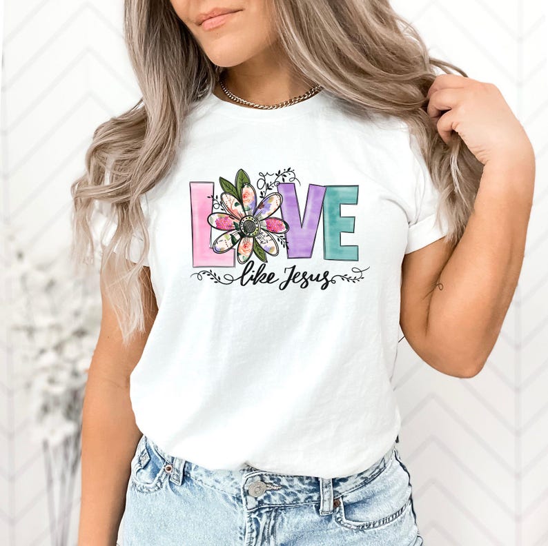 May include: White t-shirt featuring a pastel floral design with the word 'LOVE' in various pastel colors. A smaller floral design is incorporated into the letter 'O'. The phrase 'like Jesus' is written below the word 'LOVE'. The shirt is shown on a model, and the overall style is casual and religious.