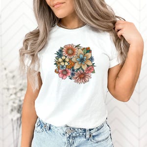 May include: White t-shirt with a colorful floral print featuring a variety of flowers in shades of pink, yellow, blue, and orange.