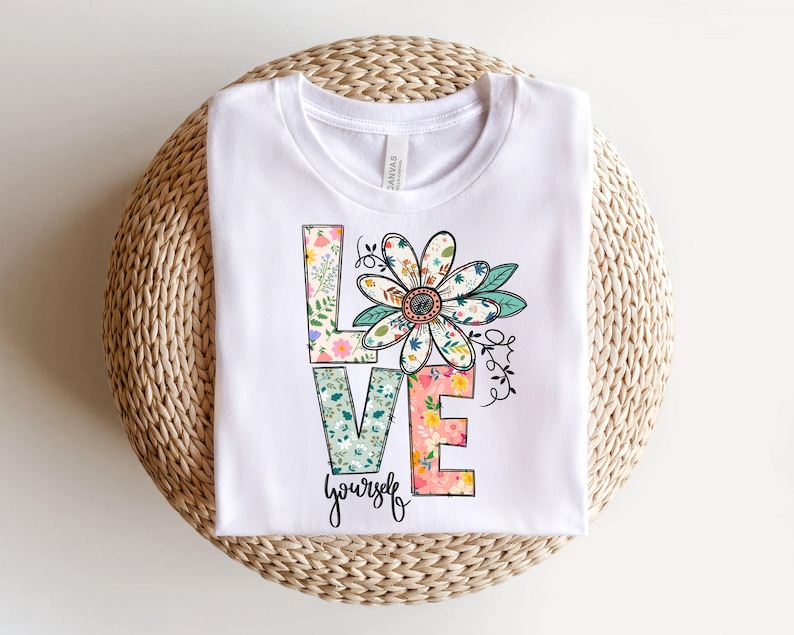 May include: White t-shirt featuring a floral design spelling out the word 'LOVE' with the phrase 'yourself' underneath. The flower is multicolored with pink, green, and blue tones. The letters are decorated with various floral patterns in pastel colors. The shirt is displayed folded on a woven surface.