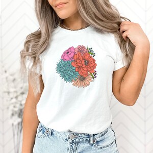 Pu&ograve; includere: Maglietta bianca con stampa di un bouquet di fiori colorati. Il bouquet comprende fiori rosa, rossi, arancioni, blu e verdi.