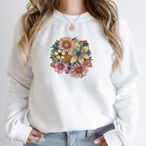 May include: White sweatshirt with a colorful floral print on the front. The flowers are in shades of pink, yellow, blue, and green.