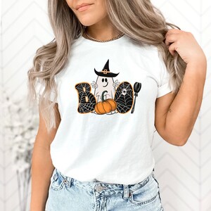 May include: White t-shirt featuring a graphic of a ghost wearing a witch's hat and holding a pumpkin. The graphic is surrounded by spiderwebs and the word "BOO" is written in black letters.