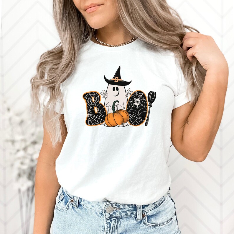 May include: White t-shirt featuring a graphic of a ghost wearing a witch's hat and holding a pumpkin. The graphic is surrounded by spiderwebs and the word "BOO" is written in black letters.