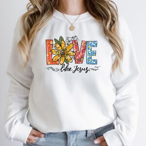 May include: White sweatshirt with a floral design that spells out "LOVE" with the words "like Jesus" underneath.