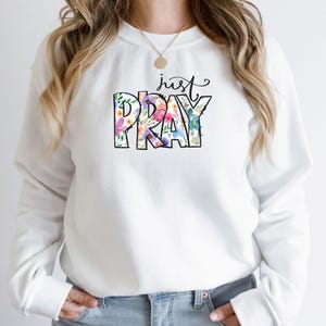 Pu&ograve; includere: Felpa bianca con un design floreale che dice "just pray" in testo nero.