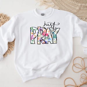Pu&ograve; includere: Felpa bianca con il testo "just pray" in lettere nere con accenti floreali.
