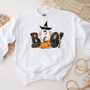 May include: White sweatshirt with a black and orange Halloween graphic. The graphic features a ghost wearing a witch hat, a pumpkin, and the word "BOO" with spiderwebs.
