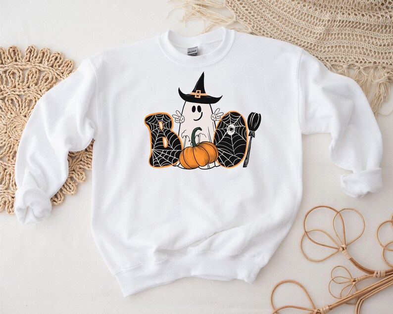 May include: White sweatshirt with a black and orange Halloween graphic. The graphic features a ghost wearing a witch hat, a pumpkin, and the word "BOO" with spiderwebs.