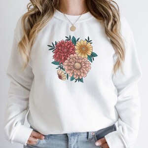 May include: White sweatshirt with a floral print featuring pink, red, and yellow flowers. The flowers are arranged in a bouquet shape and are surrounded by green leaves.