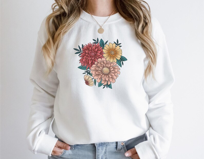 May include: White sweatshirt with a floral print featuring pink, red, and yellow flowers. The flowers are arranged in a bouquet shape and are surrounded by green leaves.