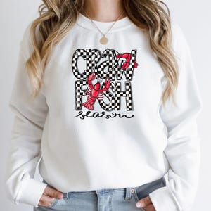 May include: White sweatshirt with a black and white checkered design that says "Crawfish Season" with a red crawfish illustration.