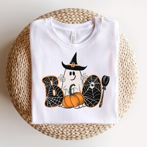 May include: White t-shirt featuring a black and orange graphic of a ghost wearing a witch's hat, a pumpkin, and the word "BOO" with spiderwebs.