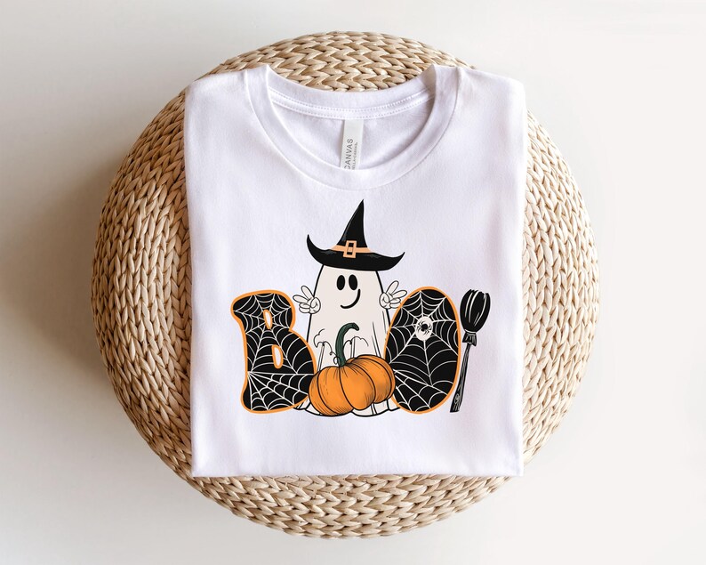 May include: White t-shirt featuring a black and orange graphic of a ghost wearing a witch's hat, a pumpkin, and the word "BOO" with spiderwebs.