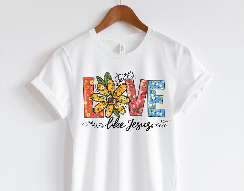 May include: White short sleeve t-shirt featuring a floral design spelling out the word LOVE.  The letters are multicolored and filled with various floral patterns. A yellow daisy is in the center of the word. Below the floral word LOVE, the text 'like Jesus' is printed in a simple, elegant font. The shirt is displayed on a wooden hanger against a white background.