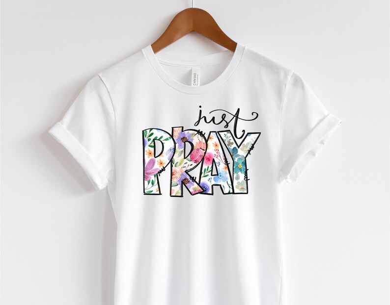 Pu&ograve; includere: Maglietta bianca con un design floreale che scrive "just pray".