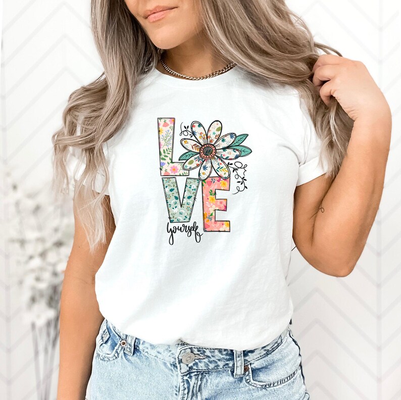 May include: White t-shirt with a floral design that spells out "LOVE" with the word "yourself" underneath.