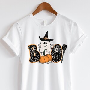 May include: White t-shirt featuring a black and white graphic of a ghost wearing a witch's hat and holding a pumpkin. The graphic also includes a spiderweb and a broom. The text "BOO" is written in black with a spiderweb design.