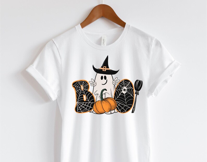 May include: White t-shirt featuring a black and white graphic of a ghost wearing a witch's hat and holding a pumpkin. The graphic also includes a spiderweb and a broom. The text "BOO" is written in black with a spiderweb design.