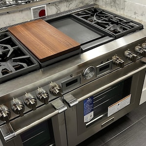 May include: A stainless steel gas range with multiple burners, a griddle, and an oven. It features a wooden chopping board, control knobs, and a digital display. The range is set against a white countertop and dark flooring.