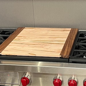 May include: A wooden chopping board with a light wood top and dark wood edging sits on top of a stainless steel hob.