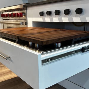 Peut inclure: Un meuble de cuisine blanc avec une planche à découper coulissante. La planche à découper est en bois foncé et se trouve au-dessus de la cuisinière. L'armoire a une poignée en acier inoxydable et des glissières de tiroir.