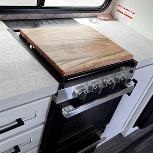 May include: A stainless steel gas range with a wooden cutting board cover. The range has four burners and an oven. The kitchen features white cabinets with black handles and a window.
