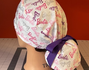 May include: White surgical scrub hat with a pink and purple "Barbie" print and a purple ribbon tie.