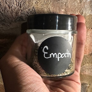 Könnte beinhalten: Ein kleines Glas mit schwarzem Deckel und einem schwarz-weißen Etikett mit der Aufschrift "Empath". Das Glas ist mit getrockneten Kräutern gefüllt.