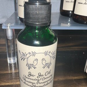 May include: A green glass bottle with a black dropper top and a brown paper label. The label has a decorative design and the text "Ju Ju Oil Uncrossing/Crossing Blessings Perseverance Shrissythecrystalindy".
