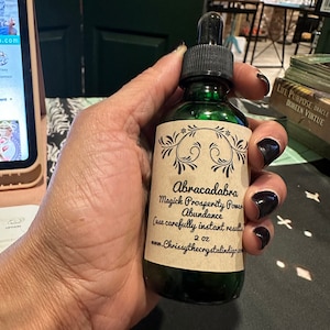 Può includere: Una bottiglia di vetro verde con un contagocce nero, tenuta in mano. L'etichetta recita "Abracadabra Magick Prosperity Power Abundance" e "2 oz". La bottiglia è probabilmente una pozione o un olio essenziale.