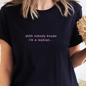 May include: Black t-shirt with the text "shhh nobody knows i'm a lesbian..." in pink lettering. The shirt has short sleeves and a crew neck. The text is centered on the chest.