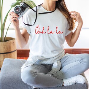 May include: A white t-shirt with the text "Ooh la la" in red cursive script. The person is wearing grey jeans and white sneakers.