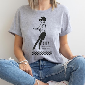 May include: A grey t-shirt featuring a black and white illustration of a woman dancing. The text "SKA THE ORIGINAL RUDE GIRL" is printed below the illustration.