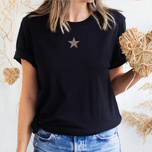May include: A black t-shirt with a gold star on the front. The t-shirt is being worn by a person and is tucked into blue jeans.