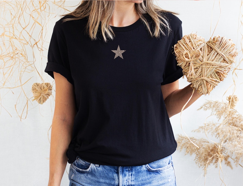 May include: A black t-shirt with a gold star on the front. The t-shirt is being worn by a person and is tucked into blue jeans.