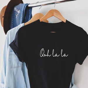 May include: Black t-shirt with white text that reads "Ooh la la" hanging on a wooden hanger.